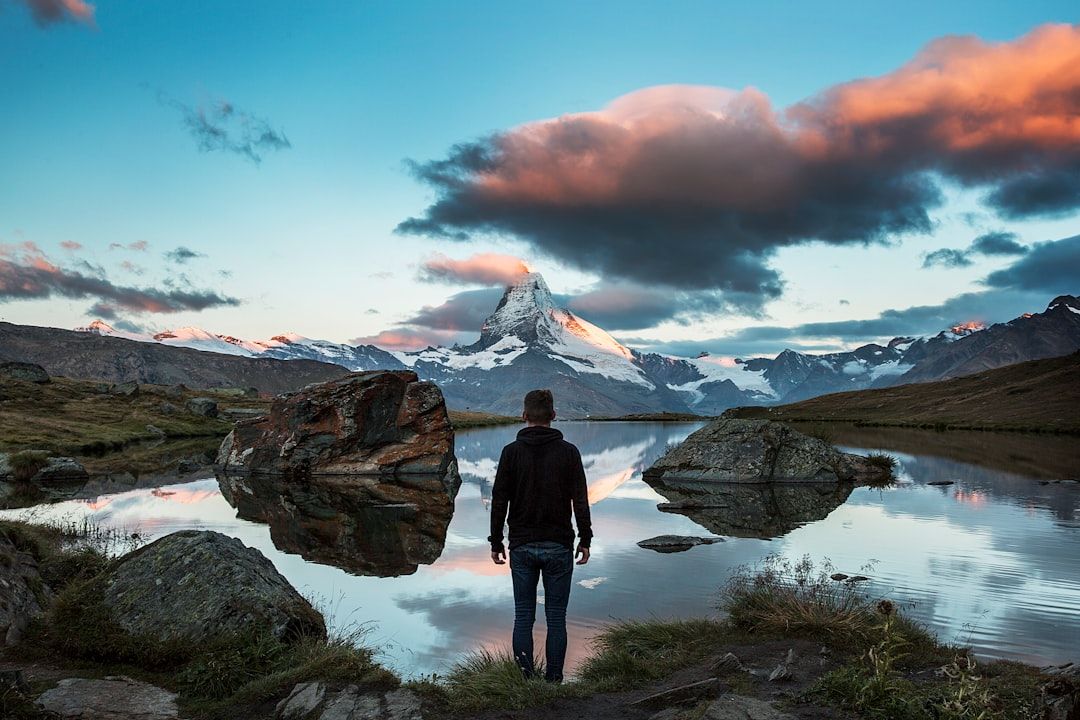 A person stands at the edge of a calm lake, facing distant snow-capped mountains under a sunset-lit sky—large rocks scattered along the shore highlight the deep human connection with nature in this tranquil scene.